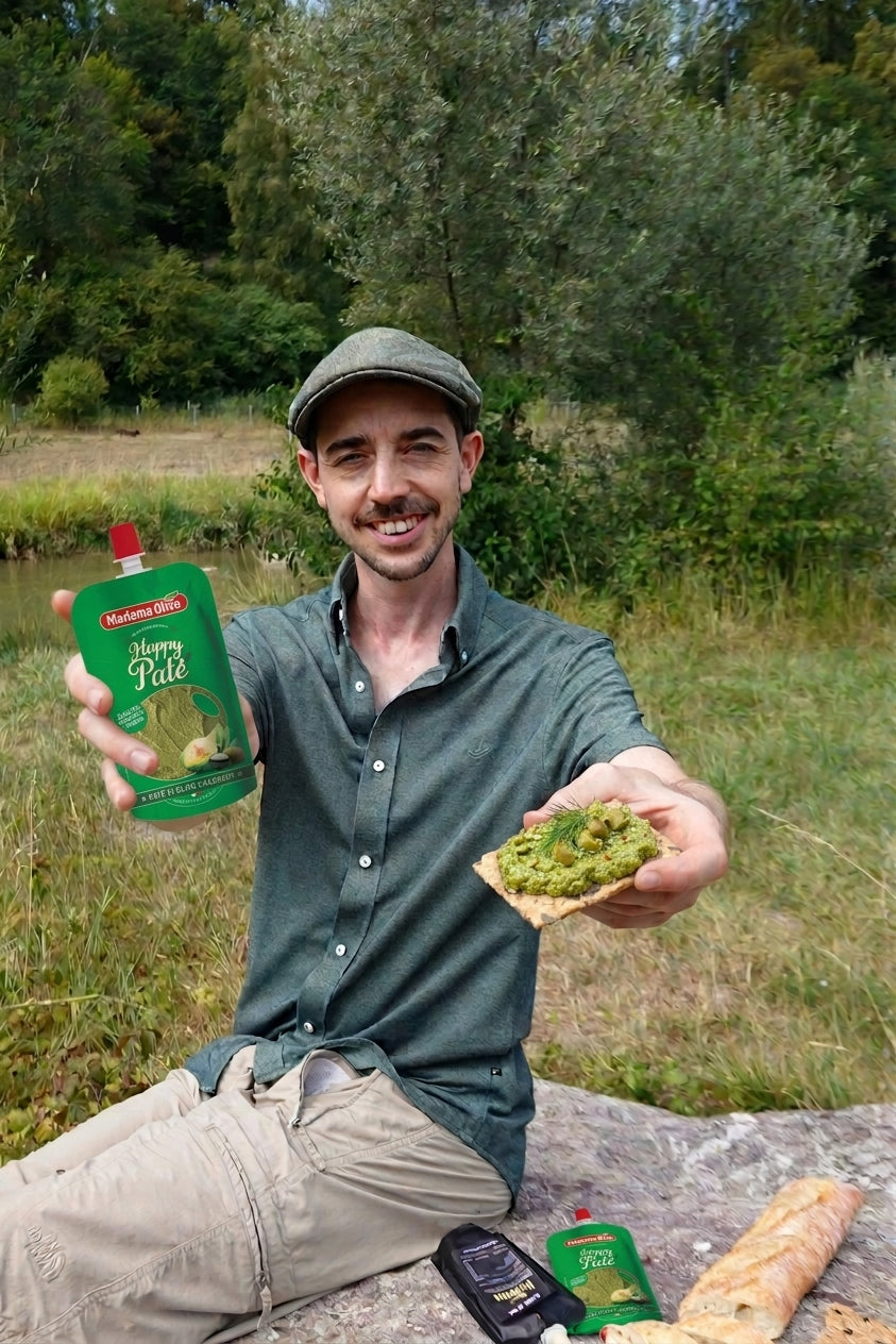 A man sitting outdoors on a stone by a grassy field holds up a green pouch of Nutrafood 'Pâté' spread and a piece of bread topped with the green spread. Trees and grass are visible in the background.